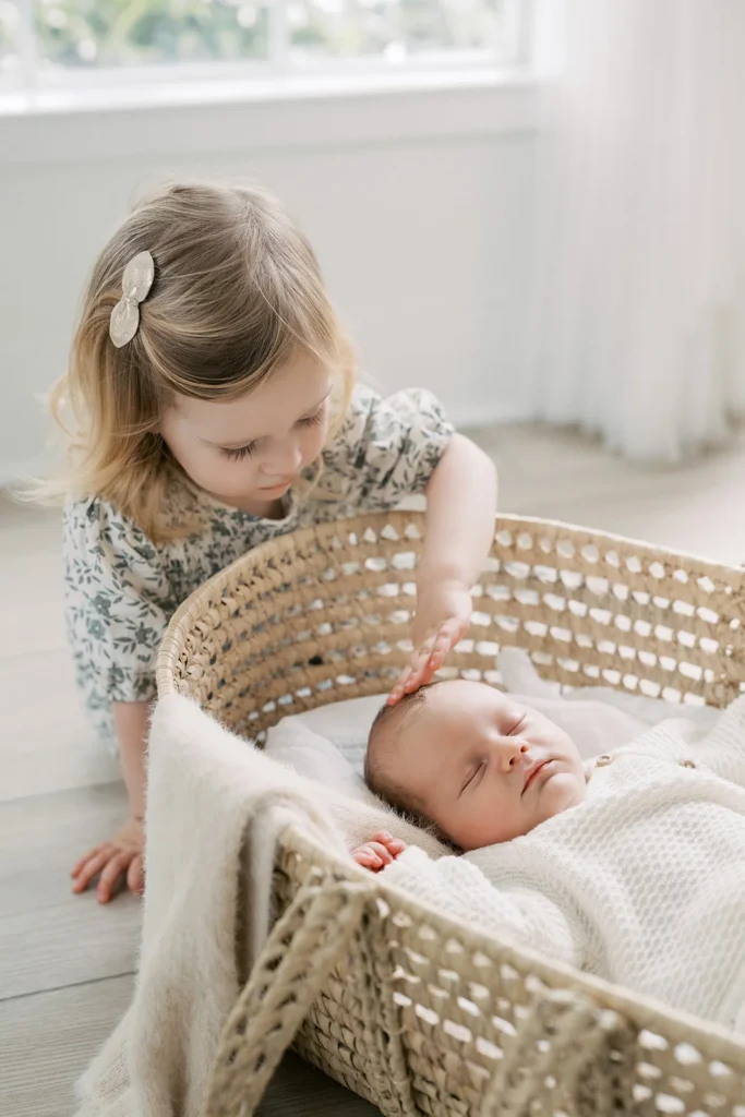 Young sibling carefully touching a sleeping newborn lying in a woven basket on the studio floor, photographed in a calm neutral Sacramento photo studio environment showcasing tender family interactions often created at best places for photoshoots in Sacramento.