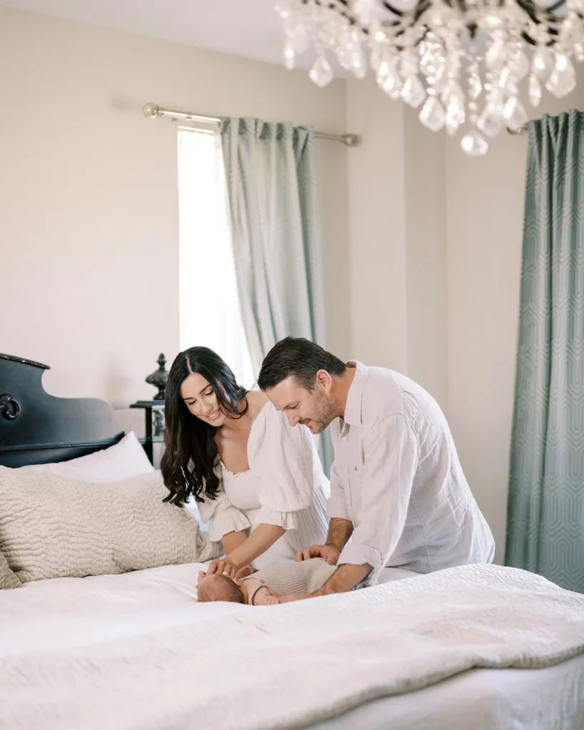 Mother and father smiling down together while adjusting their newborn baby lying on a bed beneath soft natural window light, representing authentic family interaction commonly captured at lifestyle photo studio Sacramento CA sessions and intimate photoshoot places in Sacramento homes or studios.