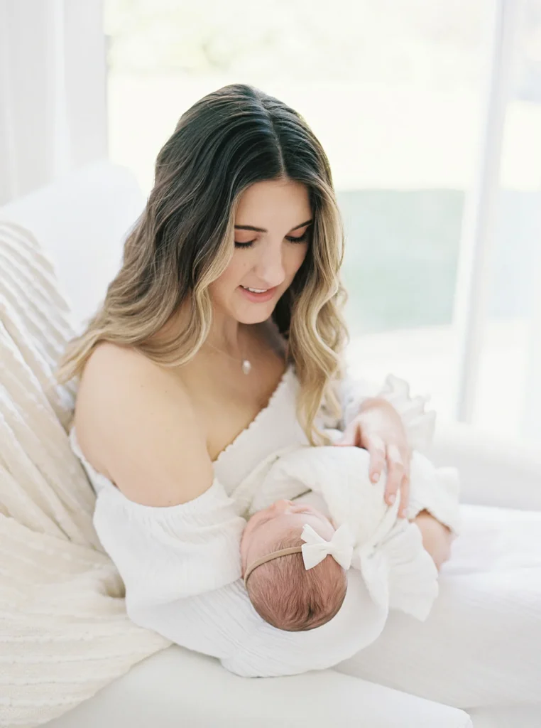 Mother seated in a bright natural-light studio gently cradling her newborn baby wrapped in a soft neutral blanket while looking down affectionately, photographed in a minimalist photo studio in Sacramento often used for timeless newborn photography and indoor photoshoot locations in Sacramento.