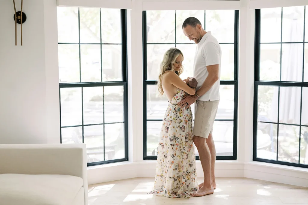 Mother and father standing together in front of tall black-framed windows while gently holding their newborn baby between them, captured in bright natural light at a modern interior often chosen as one of the best natural-light photoshoot locations in Sacramento for lifestyle newborn and family portraits.