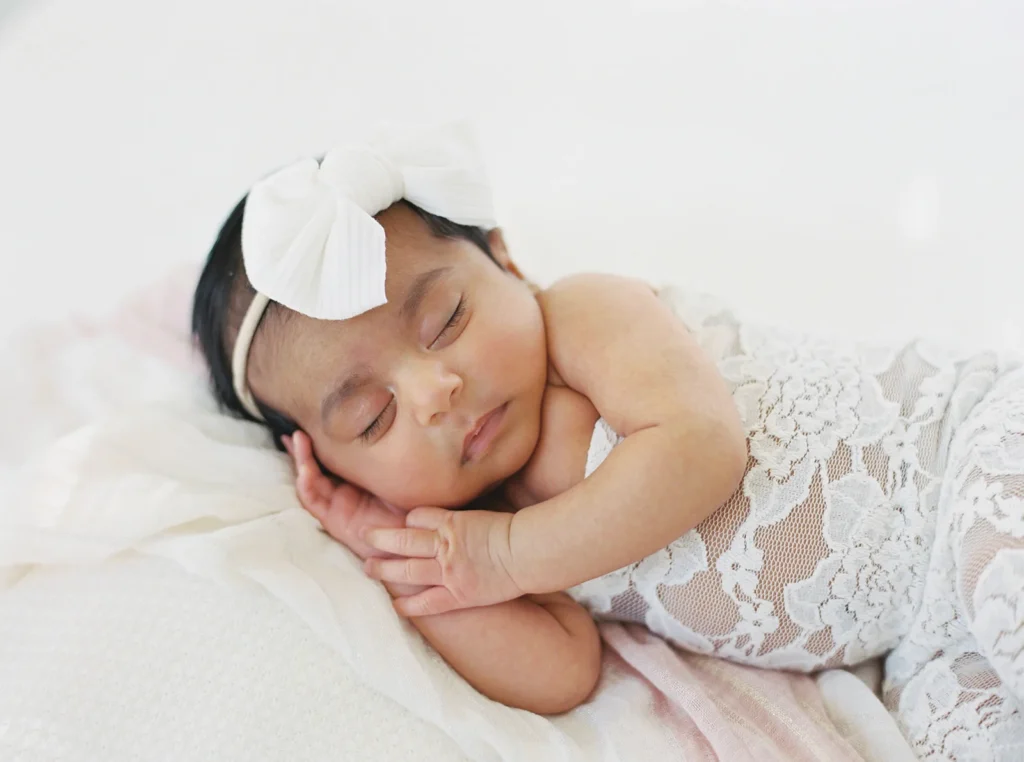 Peacefully sleeping newborn baby wearing a delicate white headband bow and lace outfit, lying on soft blankets in a minimal natural-light photo studio Sacramento CA environment, representing classic newborn portraits frequently photographed at popular photoshoot places in Sacramento studios.