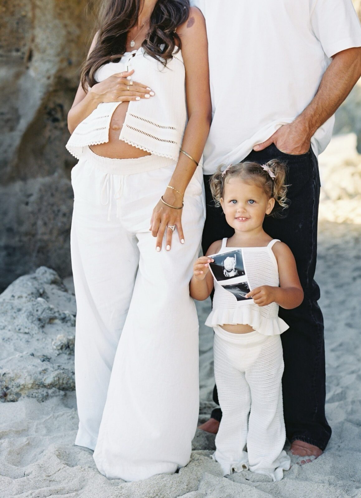 portrait of a pregnant woman with her hand on her belly, standing beside her is her husband and their younger daughter who's holding an ultrasound picture, after they followe maternity photo session tips for the best results