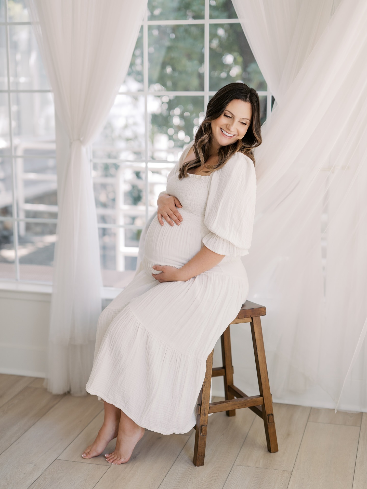 Expecting mother in a flowing white dress cradling her baby bump during a Roseville maternity photographer studio session